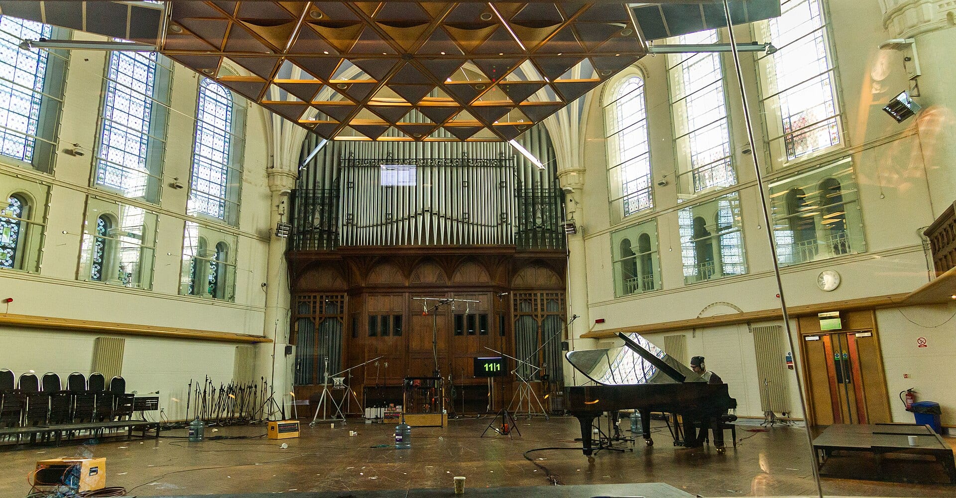 The interior of AIR Studios Lyndhurst Hall: a Victorian church nave with tall stained-glass windows and organ pipes, a suspended acoustic canopy overhead, microphone stands rigged across an empty floor and a concert grand piano to one side.