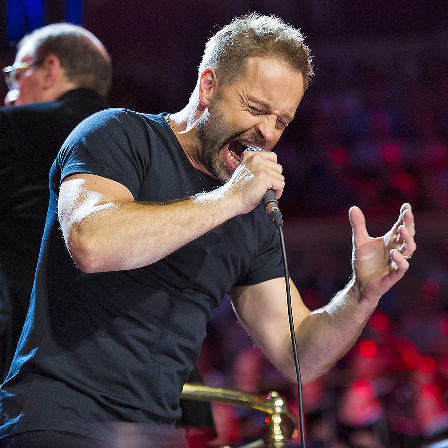 Alfie Boe singing into a handheld microphone mid-performance, eyes closed and one hand raised, stage lighting and a seated audience behind him.