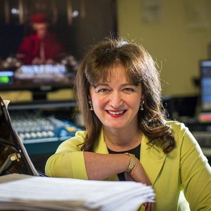 Debbie Wiseman in a recording studio control room, leaning on a piano beside a stack of manuscript paper, a mixing desk and screen behind her.