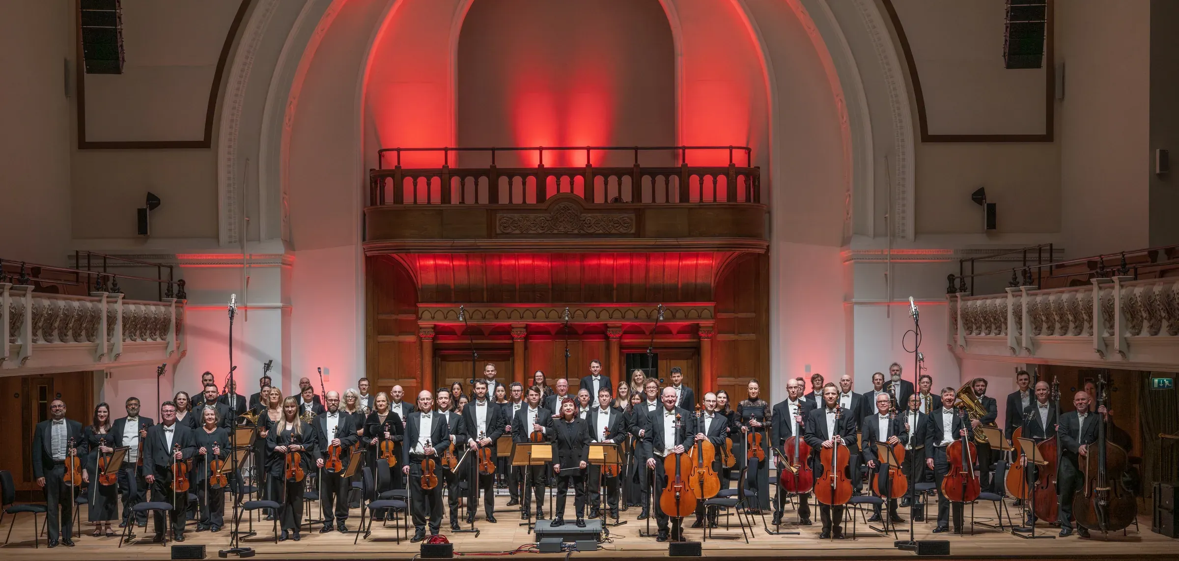The National Symphony Orchestra standing on the platform at Cadogan Hall under red light, in evening dress