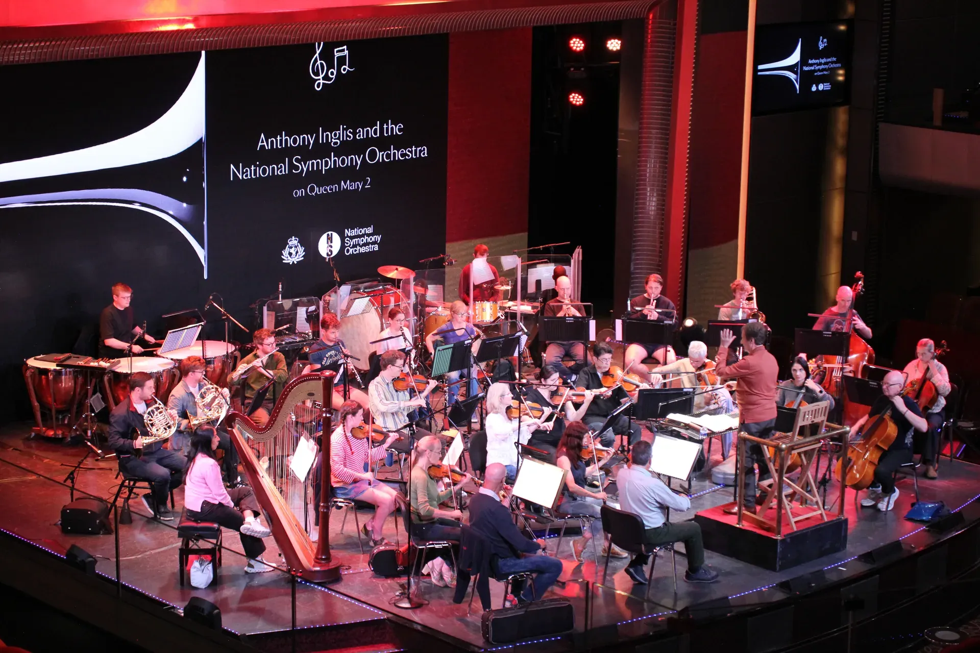 The National Symphony Orchestra rehearsing on the stage of the Queen Mary 2, the backdrop behind them reading "Anthony Inglis and the National Symphony Orchestra on Queen Mary 2".