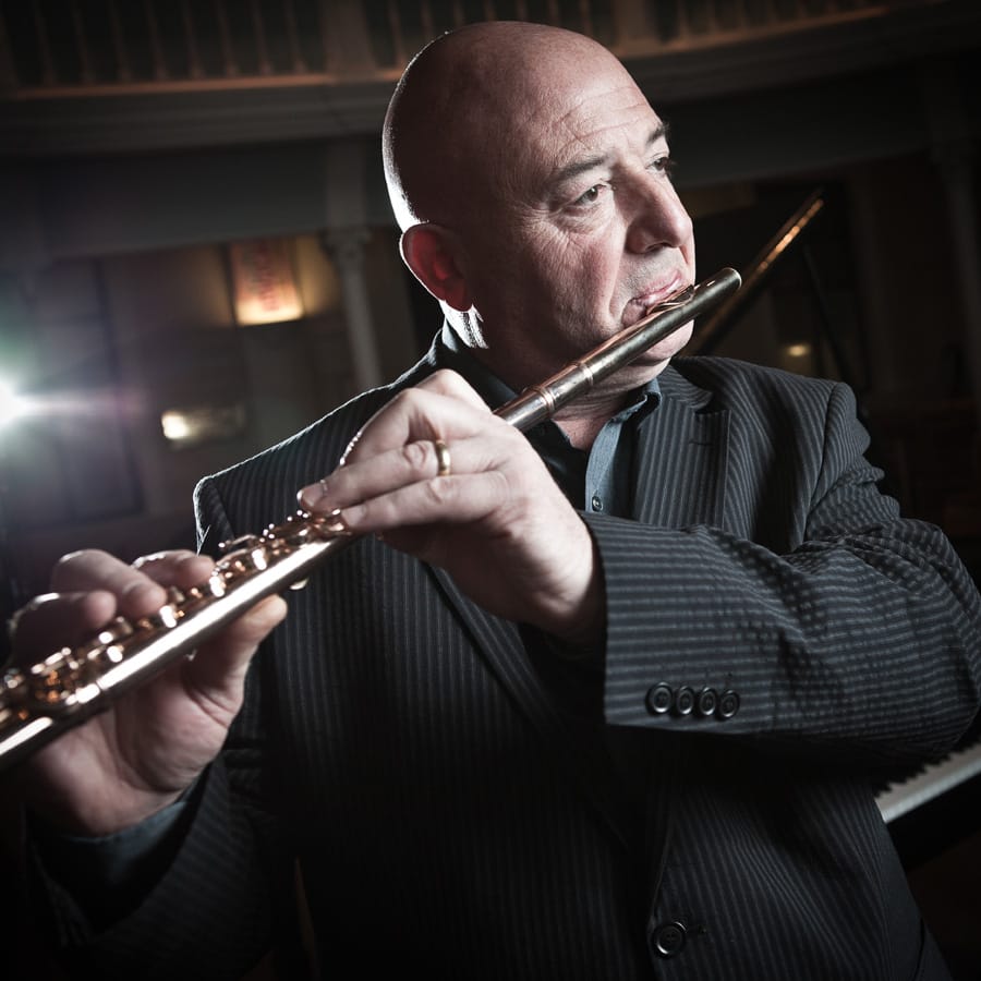 Andy Findon playing the flute in a pinstriped jacket, lit from the side in a darkened hall with a grand piano behind him.