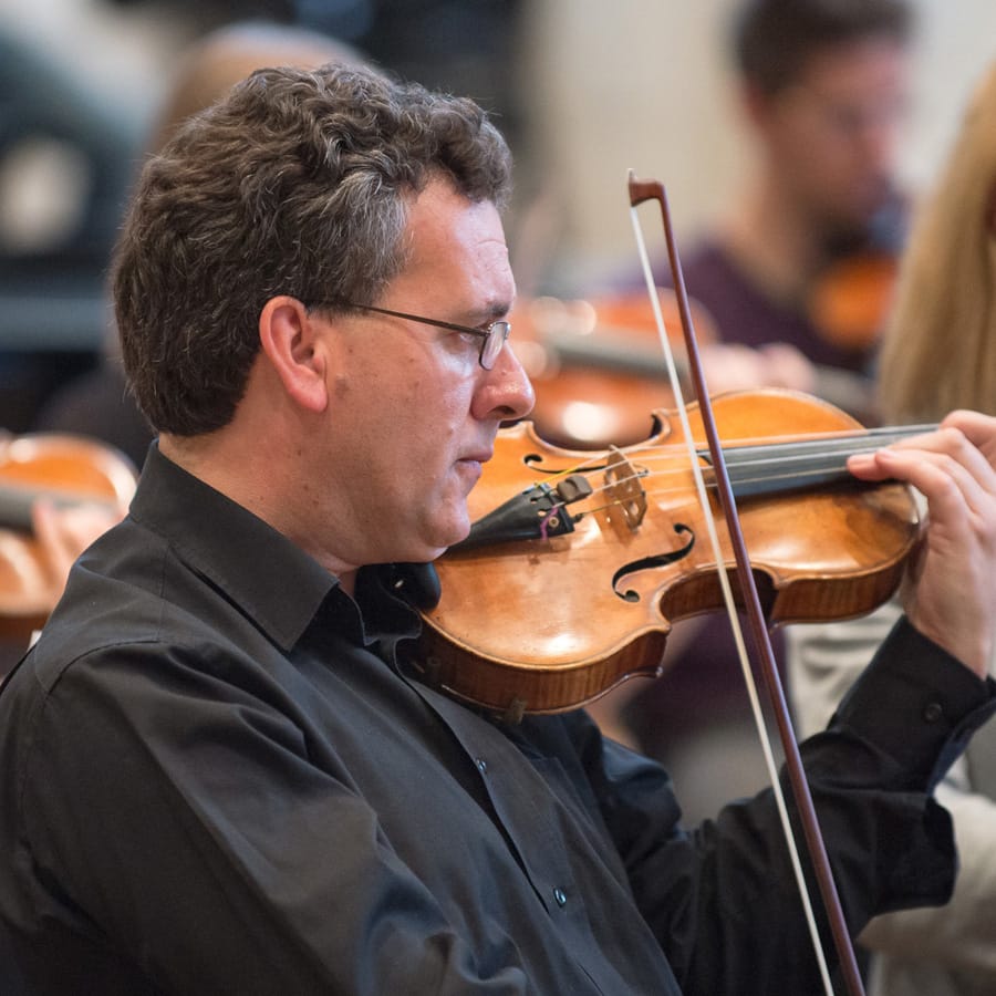 Matthew Scrivener playing the violin in rehearsal, wearing glasses and a black shirt, other string players out of focus behind him.