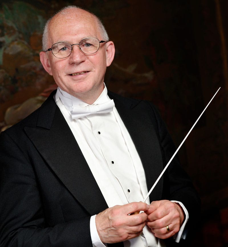 Paul Bateman in white tie and tails, holding a conductor’s baton, smiling towards the camera in front of a painted backdrop.