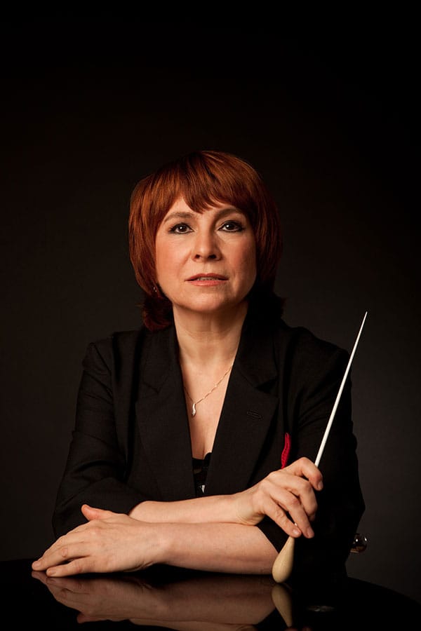 Rimma Sushanskaya in a black jacket, forearms resting on a dark reflective surface, holding a conductor’s baton upright.