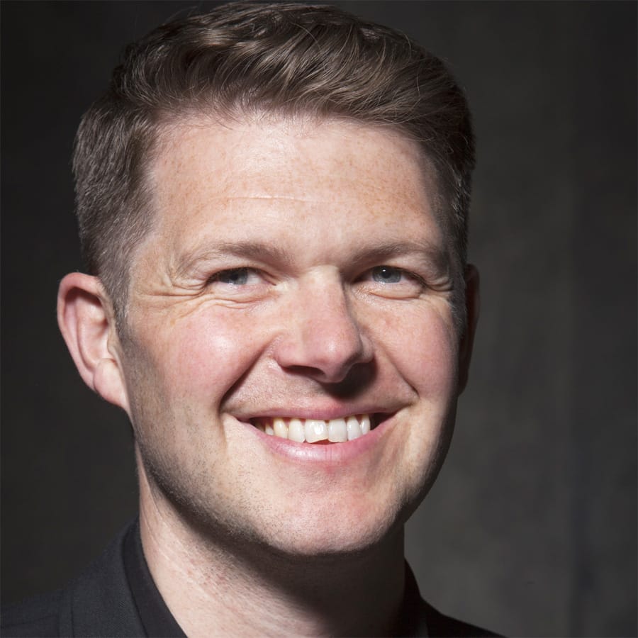 Simon Munday, head and shoulders, smiling towards the camera in a black shirt against a dark grey background.