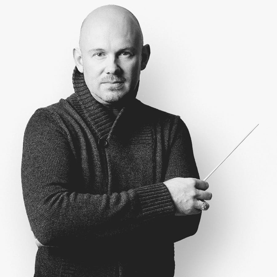Stacey Watton in a dark shawl-collar cardigan, arms folded, holding a conductor’s baton, photographed in black and white against white.